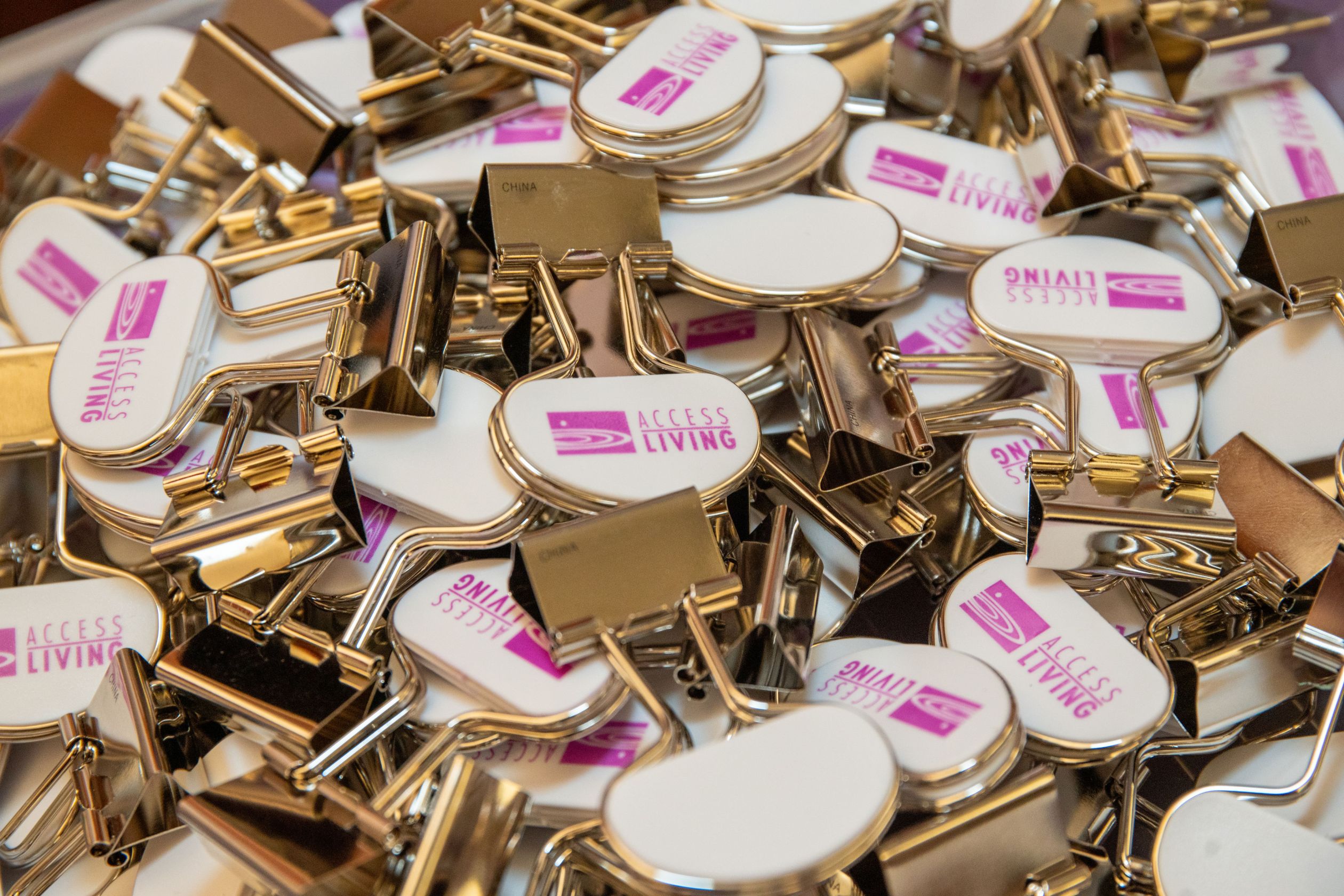 Pile of silver binder clips customized with the magenta Access Living logo on white tops.